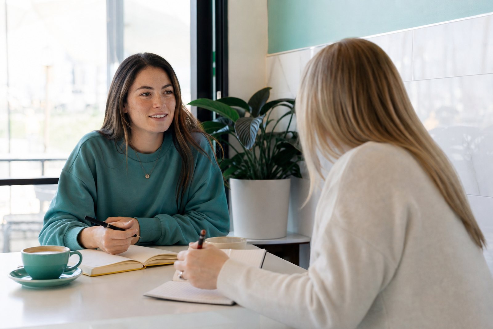 Two women meeting over coffee at a bright cafe table with open notebooks between them